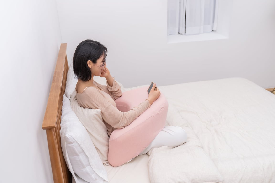 A woman resting her arms on the FIRMFLEX LevelUp Cushion while using her smartphone in bed to relieve pressure on her elbows and ulnar nerve