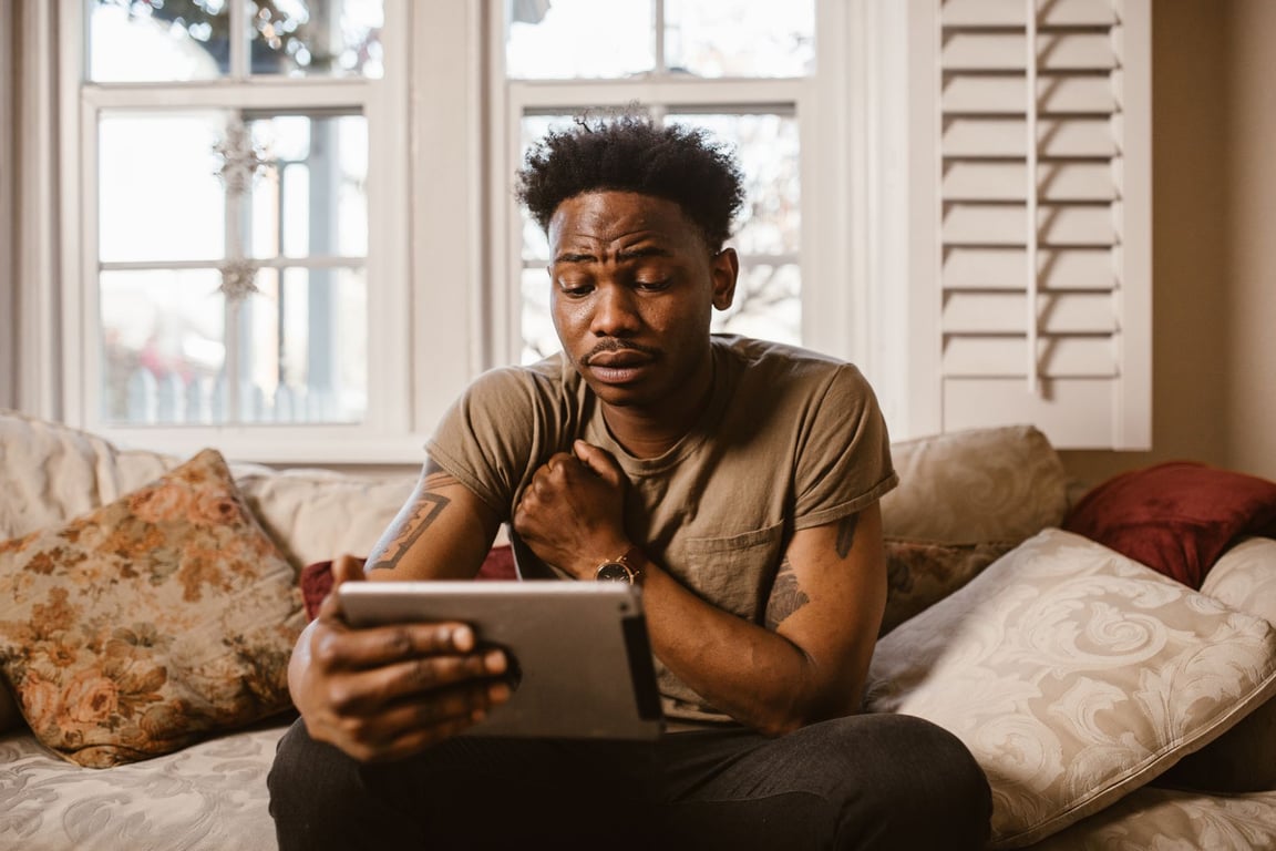 A man sitting on a sofa at home using a tablet with an emotional expression.
