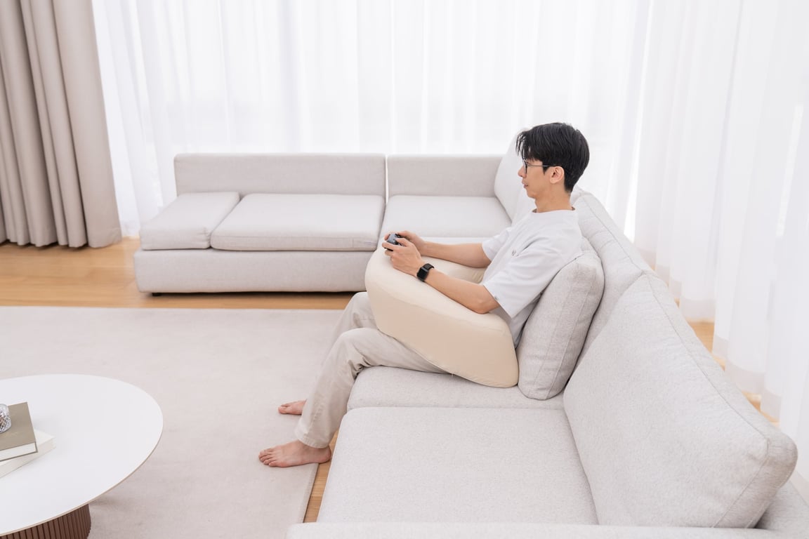 소파에 앉아 게임 리모컨을 들며 팔 아래로 베이지색 레벨업쿠션을 받치고 편안한 자세를 취하는 남성