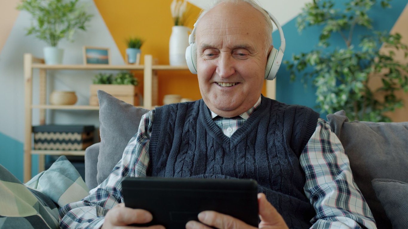 Elderly man using headphones and tablet, relaxing on couch indoors