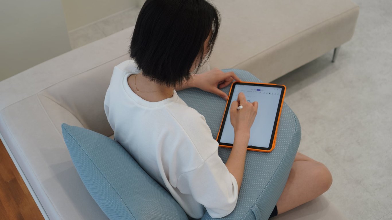 소파 앞 바닥에 앉아 팔과 무릎을 레벨업쿠션 위에 올려놓고 태블릿을 사용하는 여성, 팔 받침 구조로 어깨 긴장 완화