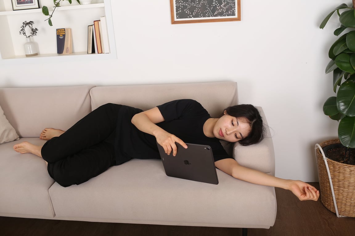 베이지 소파에 옆으로 누운 여성이 레벨업쿠션을 소파 팔걸이와 팔 사이에 끼워 팔·어깨를 받치며 태블릿을 보는 장면