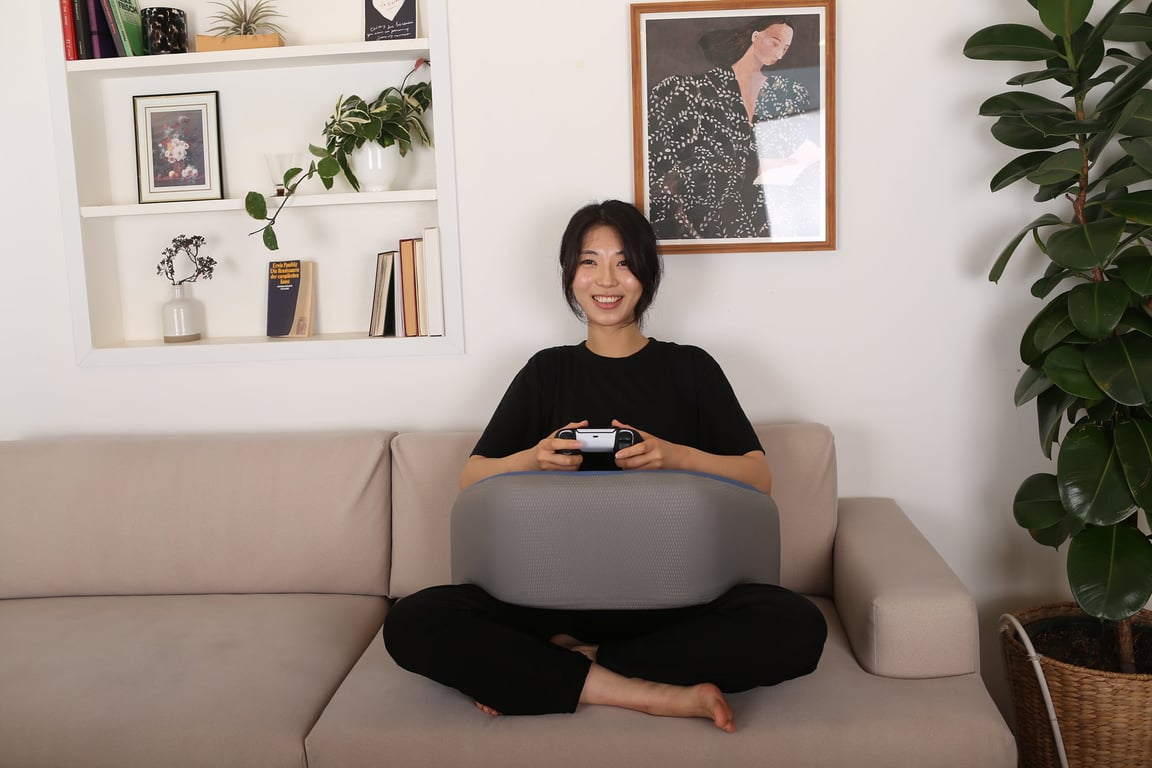 베이지 소파 위에 앉아 레벨업쿠션을 무릎에 올리고 PS5 컨트롤러를 쥐고 웃는 여성