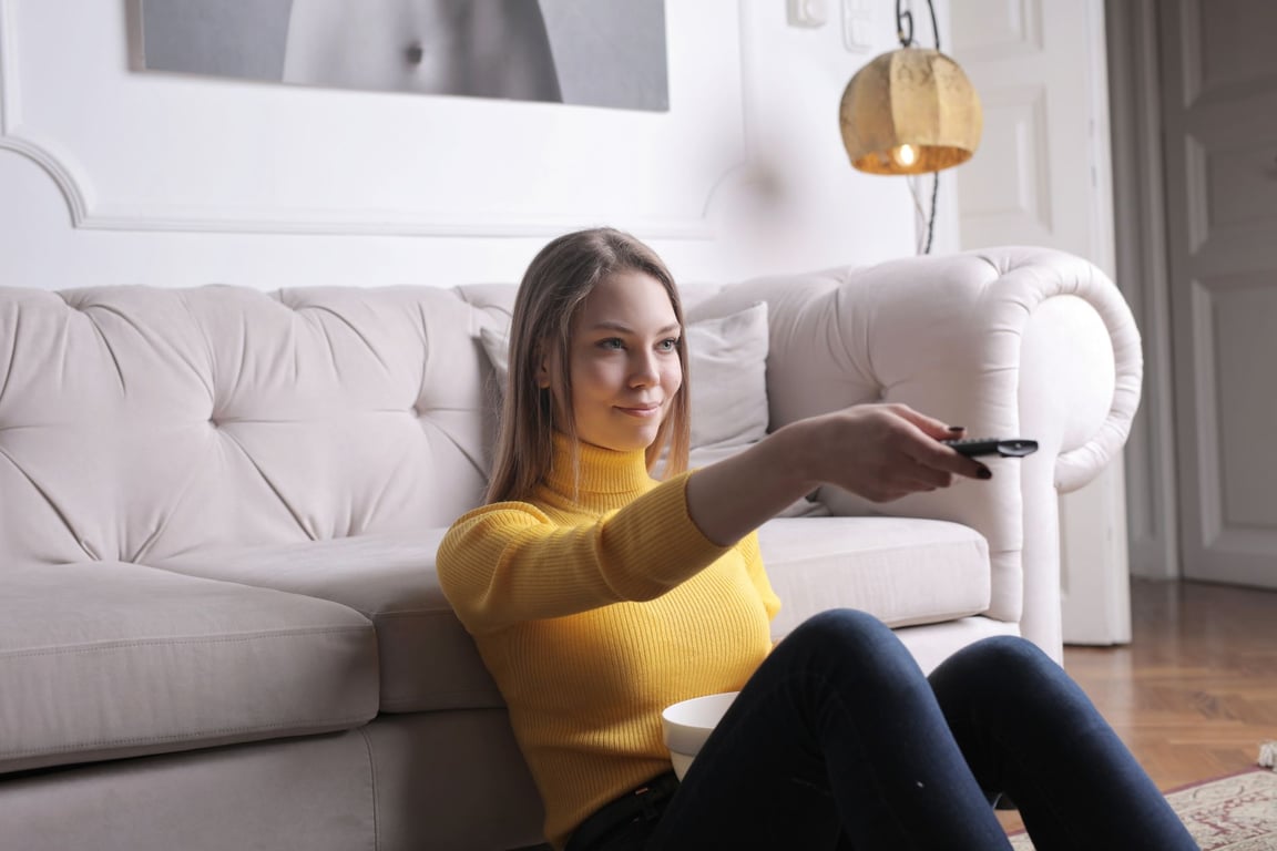 노란 스웨터를 입은 여성이 흰색 소파에 앉아 TV 리모컨을操作的하는 모습