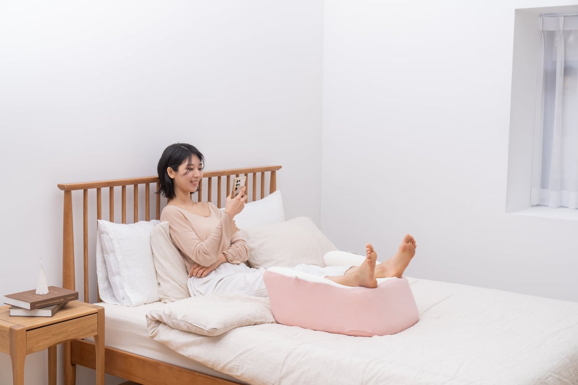 다리를 분홍색 레벨업쿠션 위에 올려놓고 침대에 누워 스마트폰을 보며 휴식하는 젊은 여성