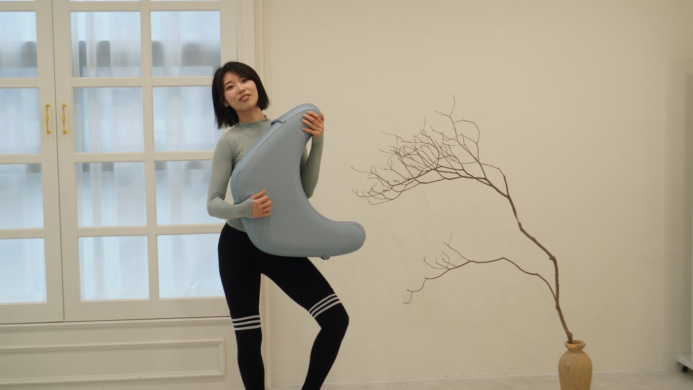 밝은 회색 레벨업쿠션을 두 손으로 안고 서 있는 단발머리 여성의 정면 샷