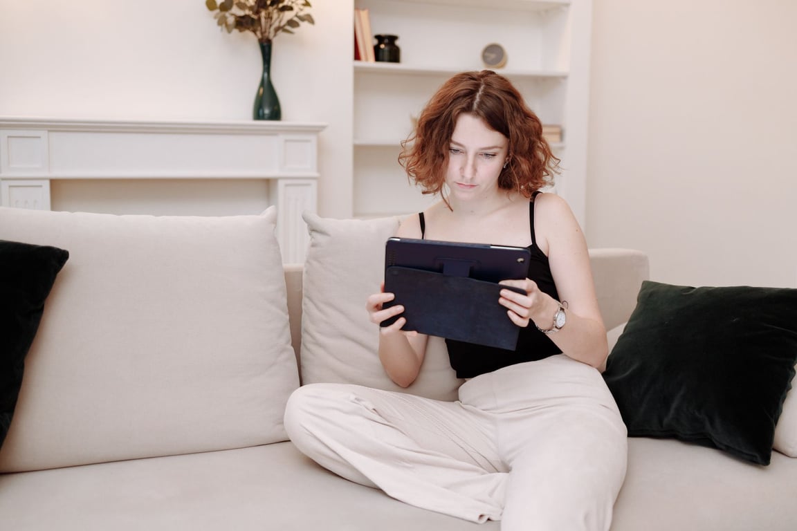 A young woman sitting on a beige couch at home, focused on using a tablet.