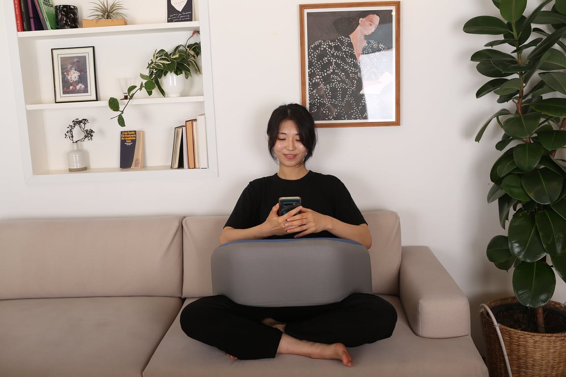소파에 앉아 양반다리 자세로 무릎 위에 레벨업쿠션을 올려놓고 두 손으로 스마트폰을 편안하게 사용하는 여성