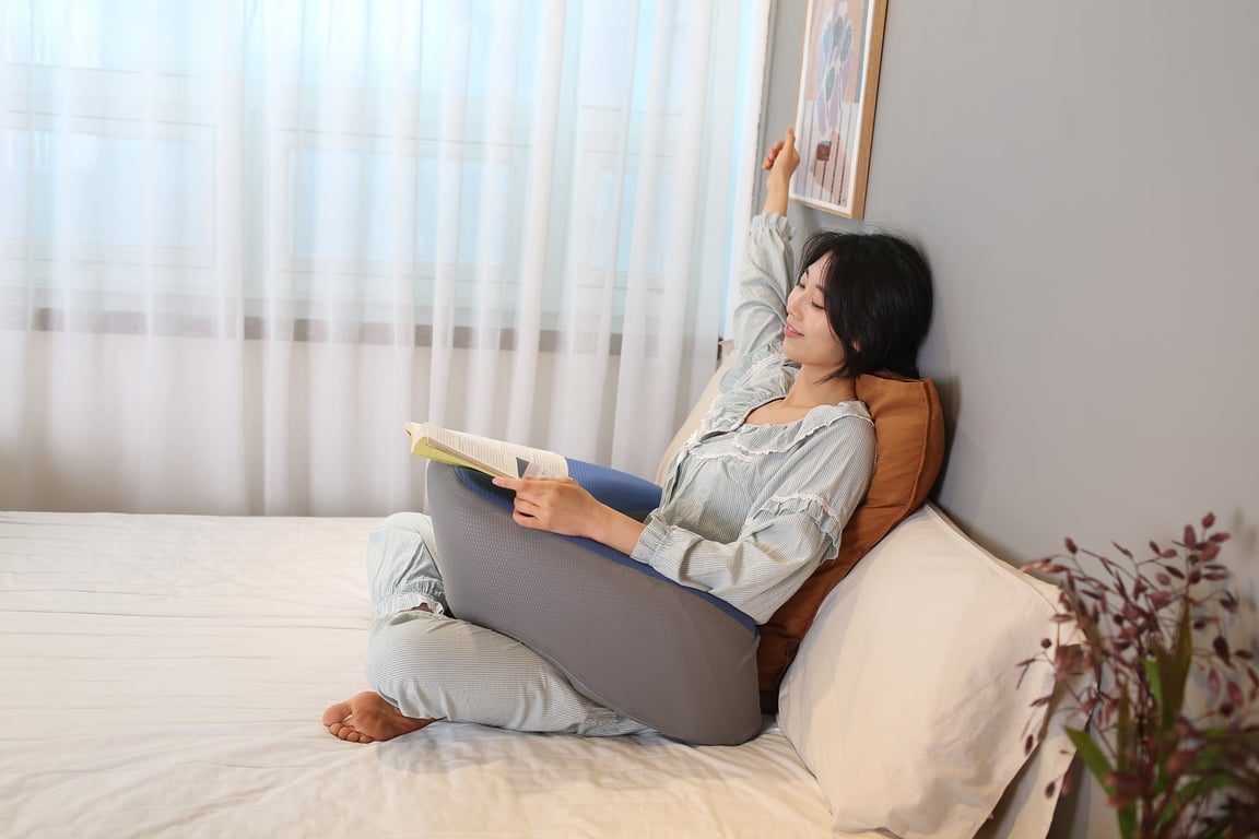 An elderly woman sitting comfortably on a bed with the FIRMFLEX LevelUp Cushion supporting her back and reading a book