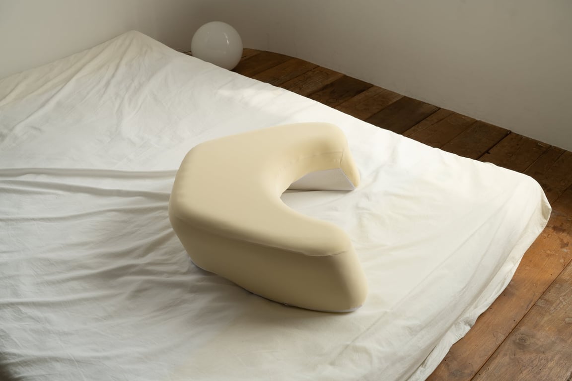 A beige FIRMFLEX LevelUp Cushion placed on a white bed sheet, highlighting its ergonomic U-shape and soft texture against a minimalist background with wooden flooring and a white lamp.