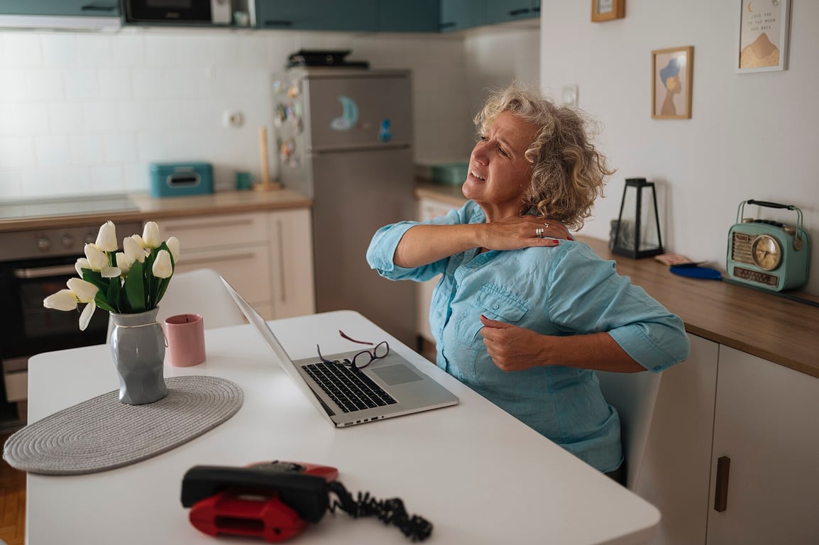 Senior woman practicing self-care and stretching in home office while working on laptop.