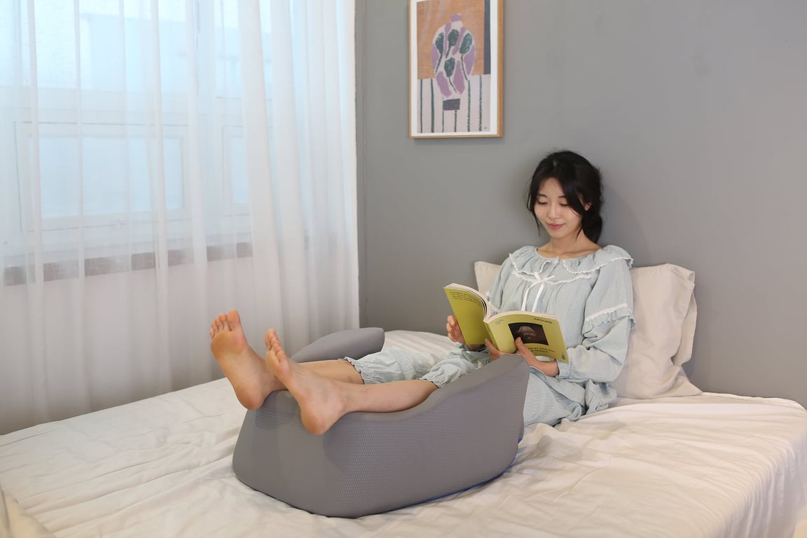 침대 위에서 무릎을 굽혀 다리를 그레이 색상의 대형 쿠션 위에 올린 채 책을 읽고 있는 여성