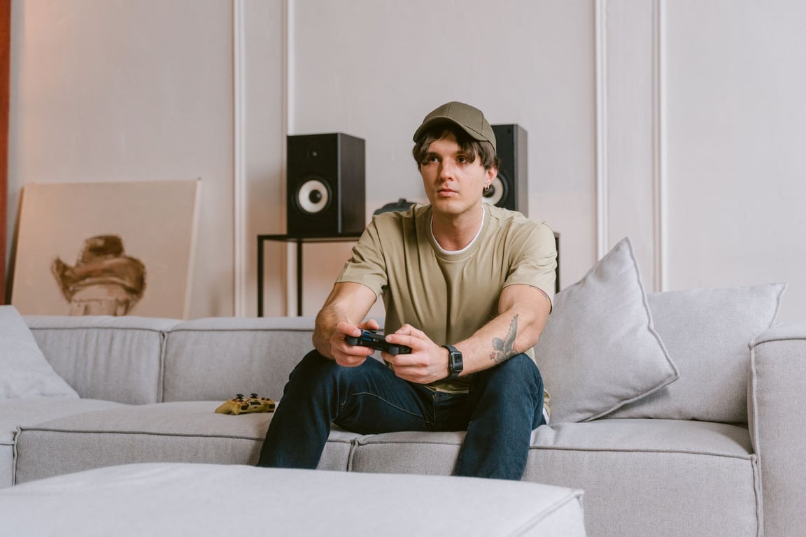 Young man wearing headphones and a cap playing video games with a controller on a living room sofa