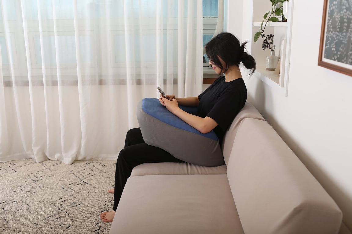 베이지 소파 모서리에 앉아 레벨업쿠션을 무릎 위에 올리고 두 팔을 쿠션에 기댄 채 스마트폰을 보는 여성