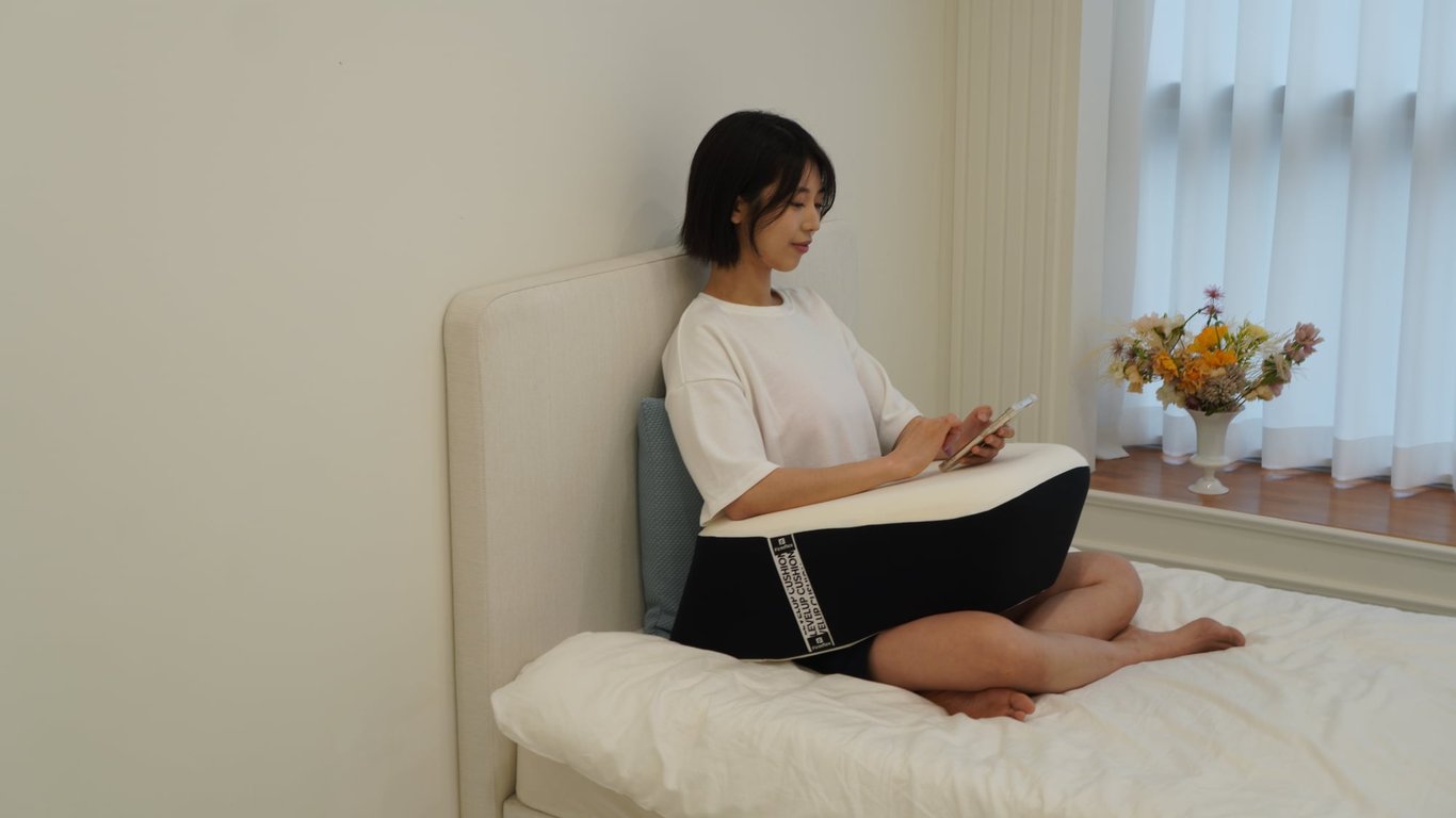 침대 위에서 가부좌 자세로 스마트폰을 사용하며 무릎 위 쿠션에 팔을 기대어 휴식하는 여성