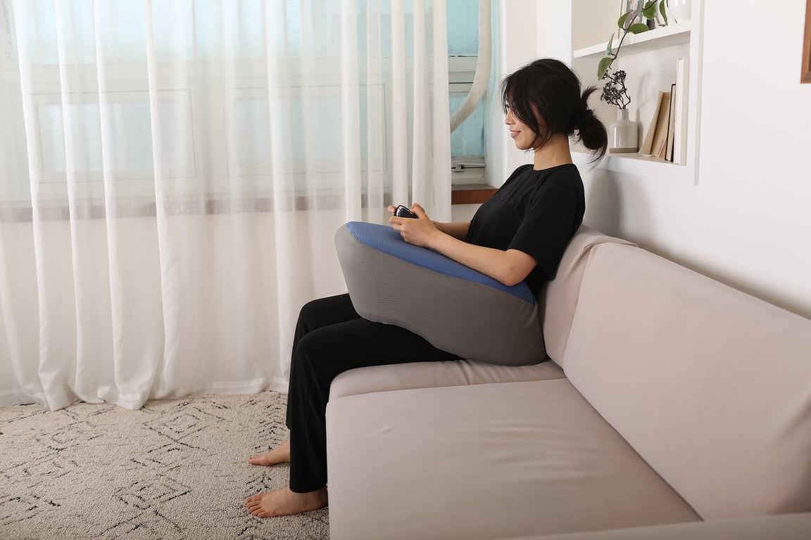 소파에 앉은 여성이 레벨업쿠션을 무릎 위에 올려놓고 양팔을 맡긴 채 게임 컨트롤러를 조작하고 있습니다.