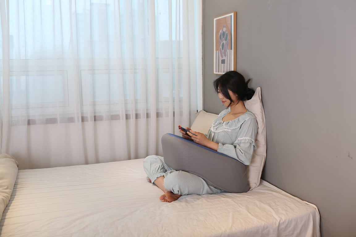 ベッド上であぐらをかいて座り、スマートフォンを使用しながらグレーのレベルアップクッションを膝の上に置いて腕を楽に支えている女性