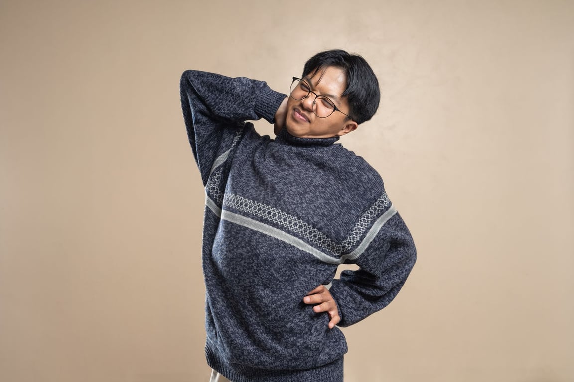 안경을 쓴 아시아 남성이 목과 어깨 통증으로 인상을 찌푸리며 스트레칭을 하고 있는 모습