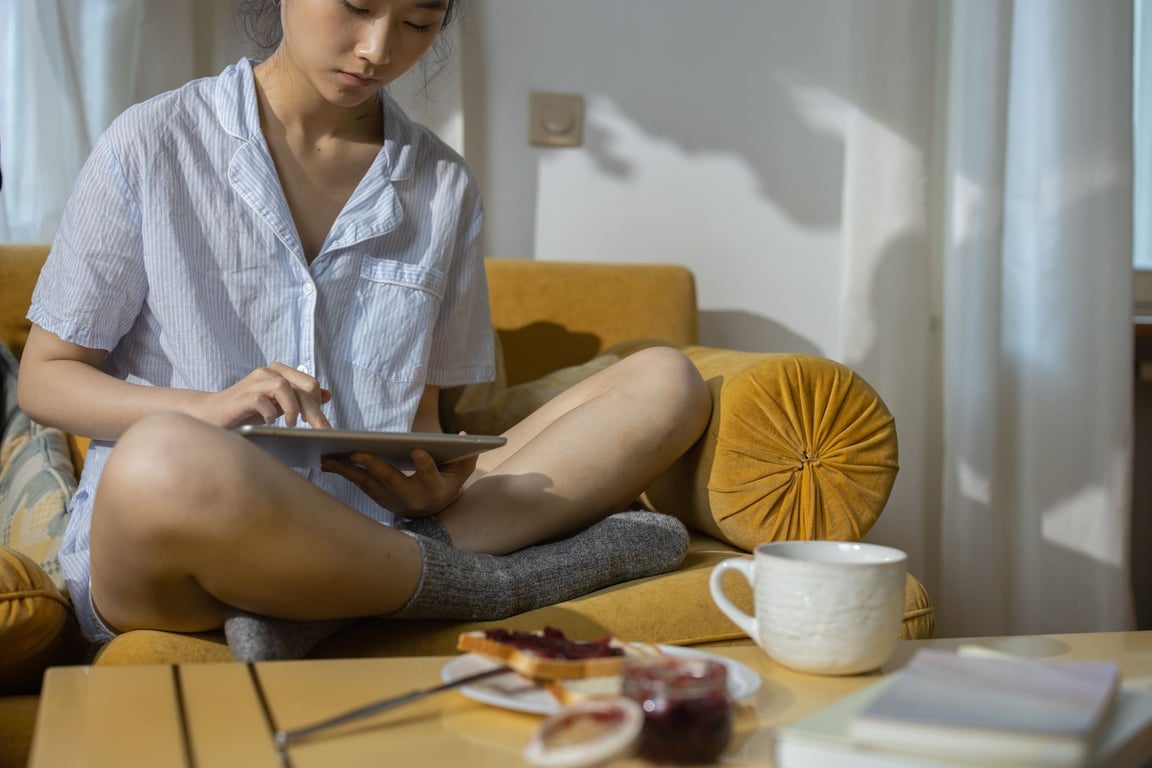 パジャマ姿でソファに座り、タブレットを操作する女性
