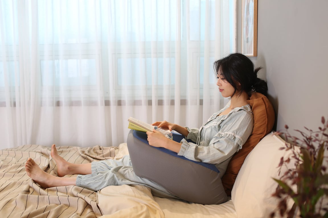 침대 위에서 책을 읽으며 무릎 위에 쿠션을 올려놓은 여성의 편안한 휴식 장면