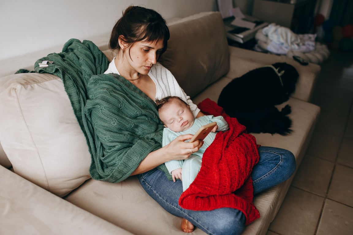 A mother resting on a sofa with her sleeping baby wrapped in blankets, creating a cozy and serene family moment at home