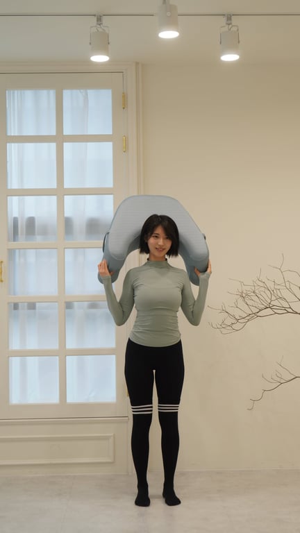 ソファのスタジオ背景でショートヘアの女性がグレーのレベルアップクッションを肩の上に載せ、製品のサイズと形状を示している