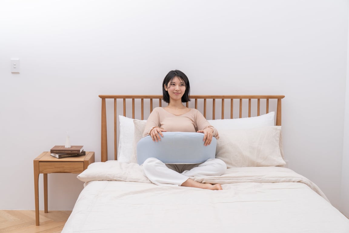 베이지색 상의와 하의를 입은 2030 여성이 나무 침대에서 다리를 굽려 앉아 연한 블루 컬러의 레벨업쿠션을 두 손으로 무릎 위에 안고 있습니다.