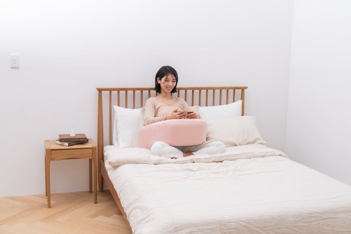 침대에 앉아 스마트폰을 보는 여성의 복부 앞에 연분홍색 레벨업쿠션이 놓여 있어 팔과 손을 부드럽게 지탱하고 있다