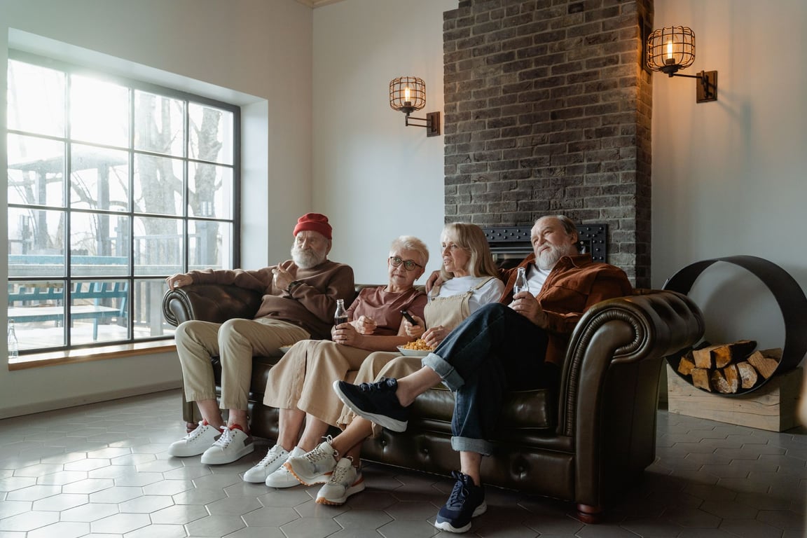 A group of senior adults relaxing comfortably indoors on a sofa, enjoying a leisurely moment together in a bright living room with a brick fireplace.