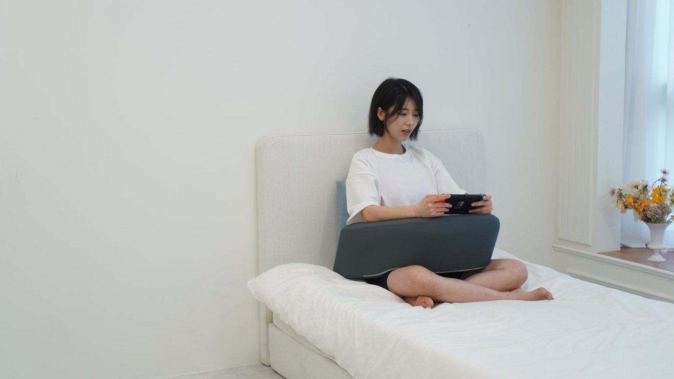 무릎 위에 레벨업쿠션을 올리고 그 위에 게임기를置いて 팔꿈치를 올린 모습. 팔의 무게를 분산시켜 어깨 결림을 완화하는 사용법을 보여줌.