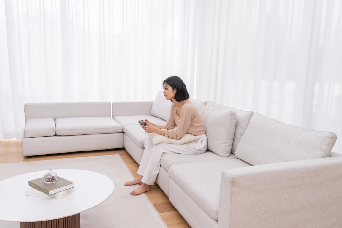 A woman sitting on a sofa playing games with the FIRMFLEX LevelUp Cushion supporting her lower back and arms