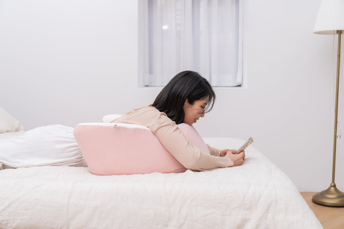 베이지색 상의를 입은 여성이 분pink색 레벨업쿠션에上半신을 기대어 침대에俯卧하며 스마트폰을 시청하고 있다
