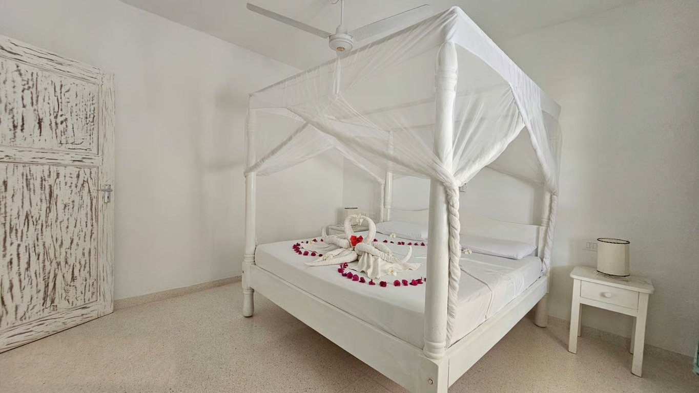 White four-poster bed beneath a mosquito net with a towel arrangement and red petals