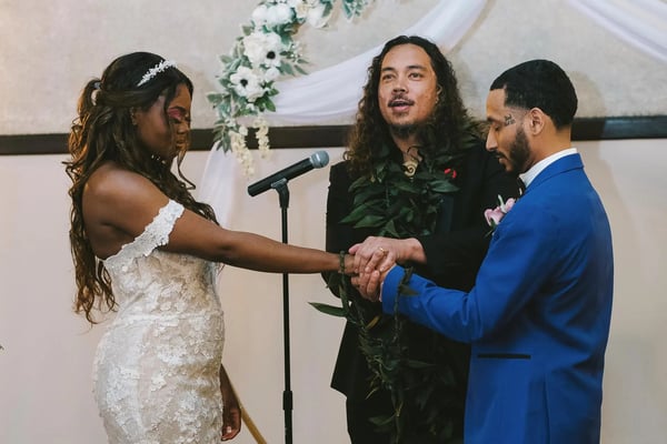 Officiant in a ti leaf lei binding a couple's joined hands with ti leaves in front of a draped floral backdrop, Hawaiian Vow Renewal Ceremony