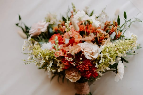 Loose bridal bouquet of peach roses, coral and red dahlias, white daisies and trailing green sprigs lying on a pale surface