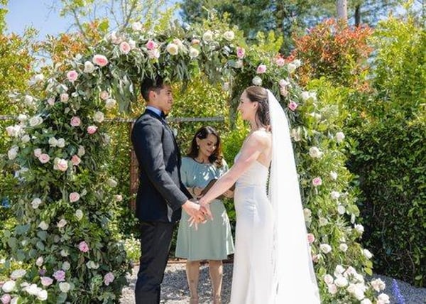 Couple holding hands under a rose-covered arch in a sunny garden while an officiant in a sage dress reads the ceremony