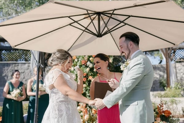 Officiant in a pink dress laughing with a bride and groom under a large patio umbrella, bridesmaids in green dresses behind them