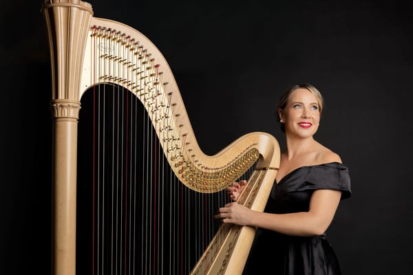 Harpist in a black off-shoulder gown smiling beside a gold concert harp against a dark backdrop, a wedding ceremony harp in San Francisco