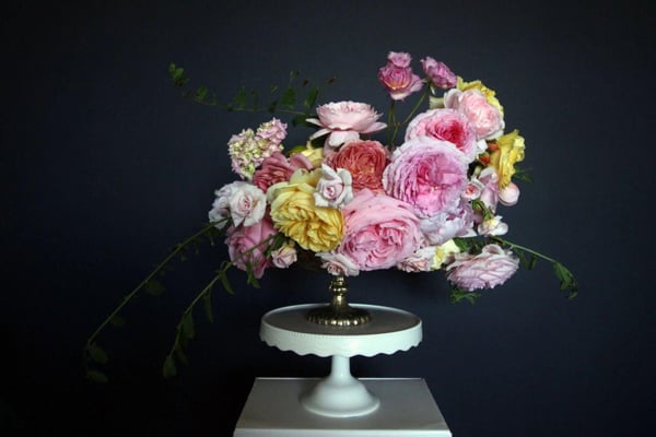 Full centerpiece of pink, peach, yellow and lavender garden roses with trailing greenery on a white scalloped cake stand against a dark backdrop
