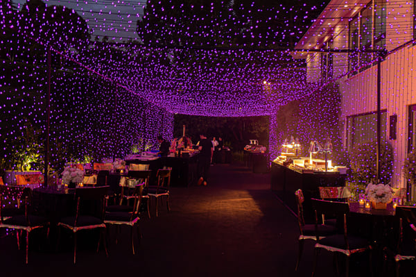 Outdoor courtyard under a canopy of purple string lights, with dining chairs, white flowers and a lit buffet line