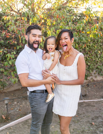 Parents and a toddler in a white dress sticking out their tongues together in front of an ivy-covered wall