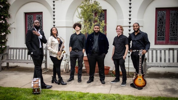 Six band members in black posing in a courtyard, one holding a saxophone and one a sunburst electric guitar, with a snare drum on the path