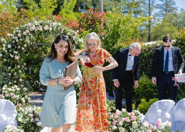 Officiant carrying a leather folder down a garden aisle beside a woman in an orange floral dress, with rose arches and white chairs