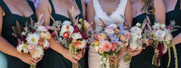 Bride and four bridesmaids in dark green dresses, shown from the shoulders down, holding bouquets of pink, peach and white flowers with dried grasses