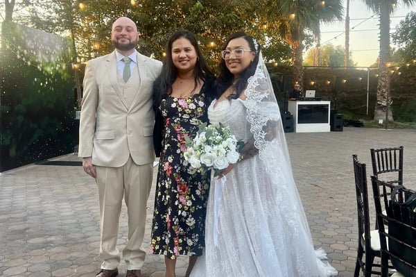 Bilingual wedding officiant from El Sobrante in a floral dress standing between a groom in a cream suit and a bride holding white roses
