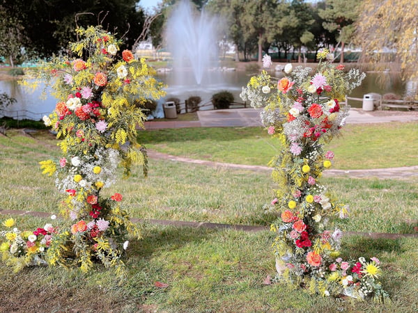 Two tall flower columns of yellow mimosa with orange, pink and red blooms standing on a lawn beside a pond with a fountain