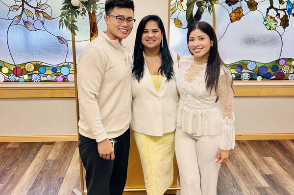 Three people smiling before a stained-glass wall and floral arch: a man in a cream sweater, a woman in a white blazer and a woman in white lace