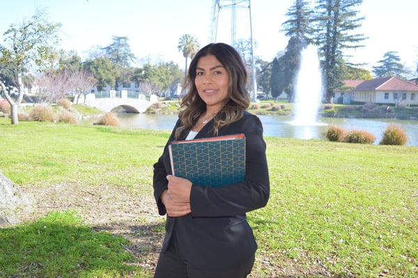 Officiant in a black suit holding a teal ceremony book in a park with a pond, a footbridge and a fountain behind her