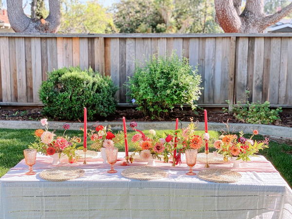 Backyard table with a white cloth, red taper candles, pink glasses, woven placemats and bud vases of orange and pink dahlias and zinnias