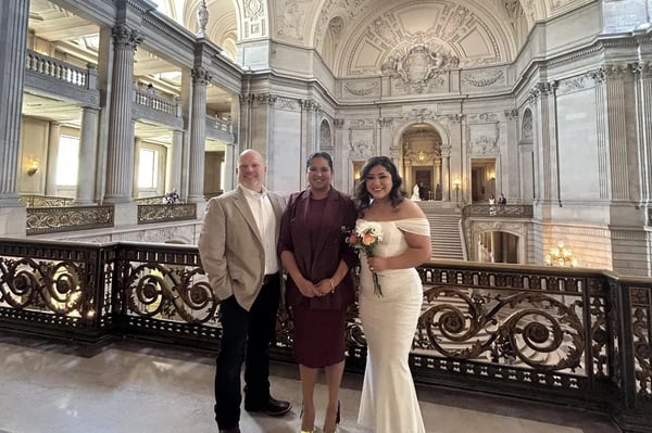 Three people posing at an ornate iron railing in a grand marble hall, including a bride in an off-shoulder white dress holding flowers